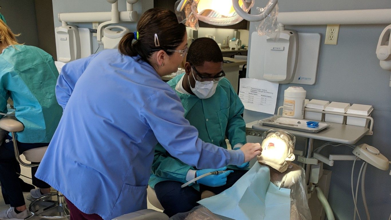 Two dental assistant students in scrubs practice a procedure under a dental exam light in a clinical lab setting