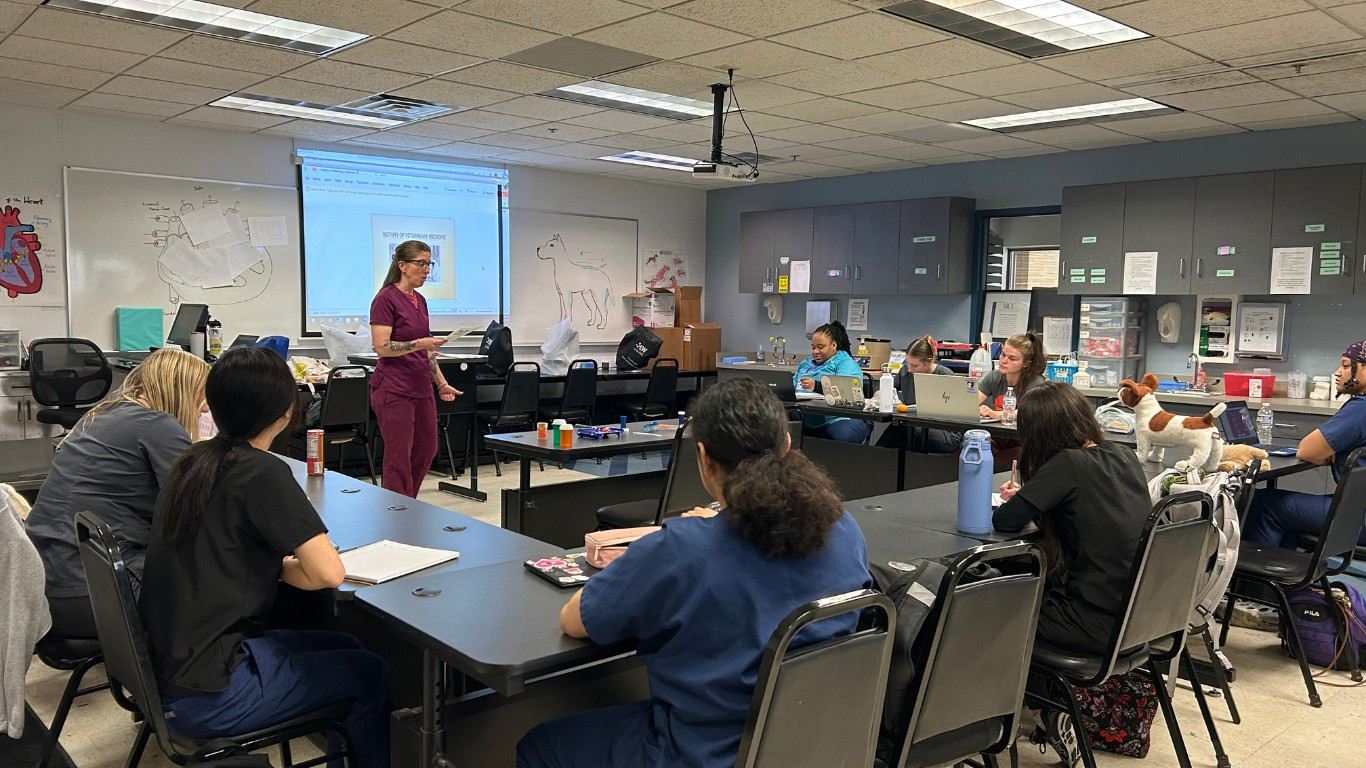 An instructor in maroon scrubs leads a veterinary assistant class in front of a projector screen. Students in scrubs sit at tables in a classroom.